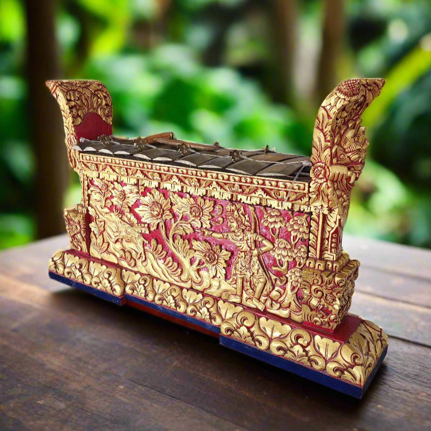 An authentic Balinese gamelan with bronze keys and a red lacquer finish, adorned with gold leaf embellishments and intricate carvings, placed on a wooden surface with a lush green background.
