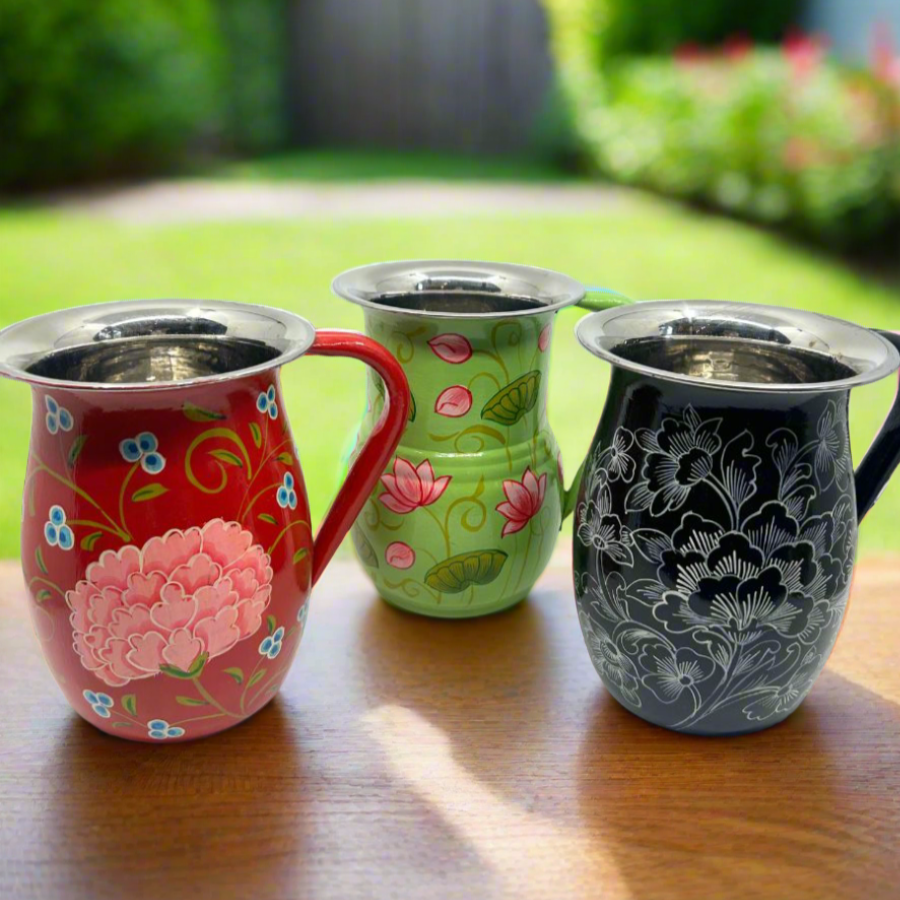 Three stainless steel pitchers with hand-painted floral and geometric patterns, displayed on a wooden surface outdoors.