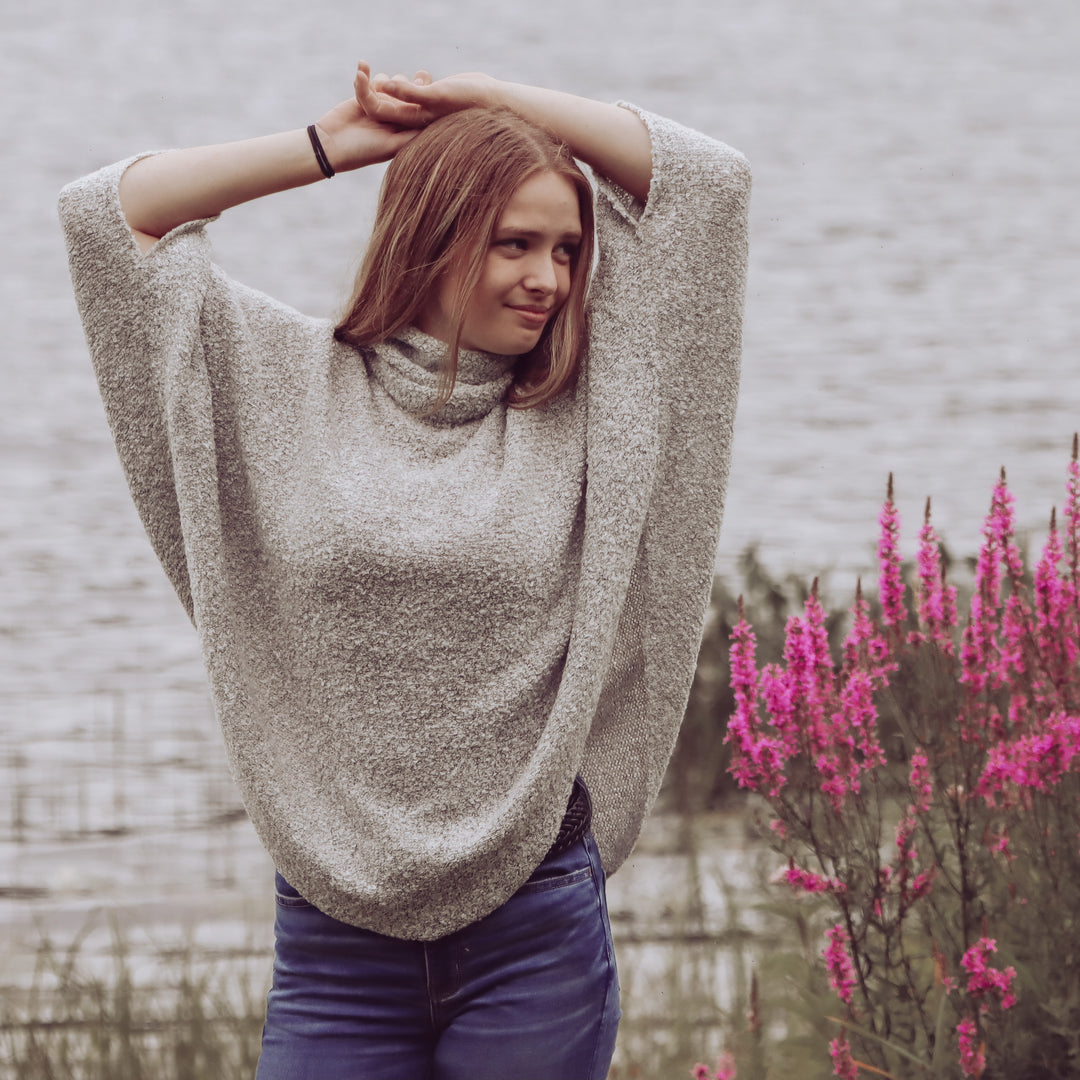 A woman standing by a body of water, wearing a grey cowl neck poncho with long sleeves, paired with blue jeans. The background includes pink flowers and a field.
