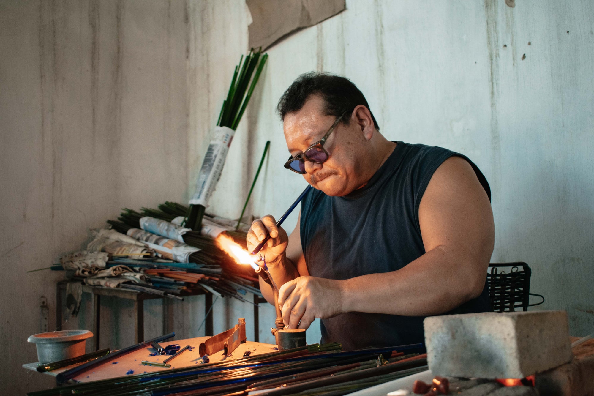 A male, Mexican national in a blue tank top with protective glasses on blows a large flaming on a workdesk to heat up a piece of pigmented 